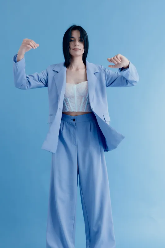 Young woman in pastel blue suit poses gracefully against a matching blue background, wearing a white lace top and showcasing modern minimalist fashion style.