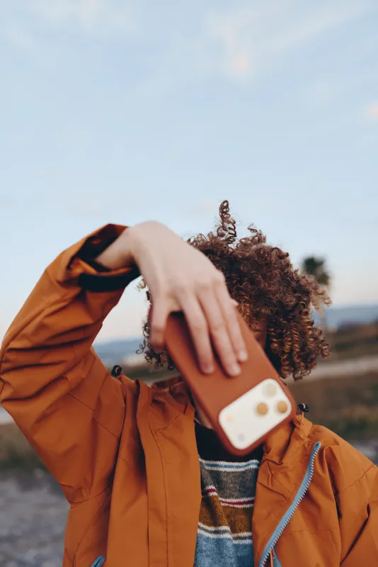 Woman lifestyle curly hair outdoors, closeup of hand holding phone, orange jacket and scarf, blurred sunset sky and horizon background, candid travel moment, soft warm, natural light