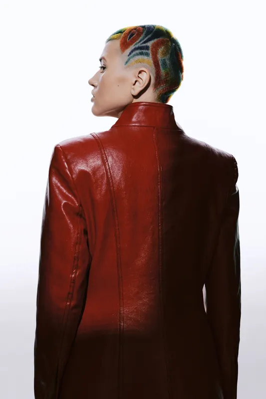 Woman with colorful patterned shaved hair wearing a red leather jacket, seen from the back against a bright white background, showcasing bold modern fashion style.