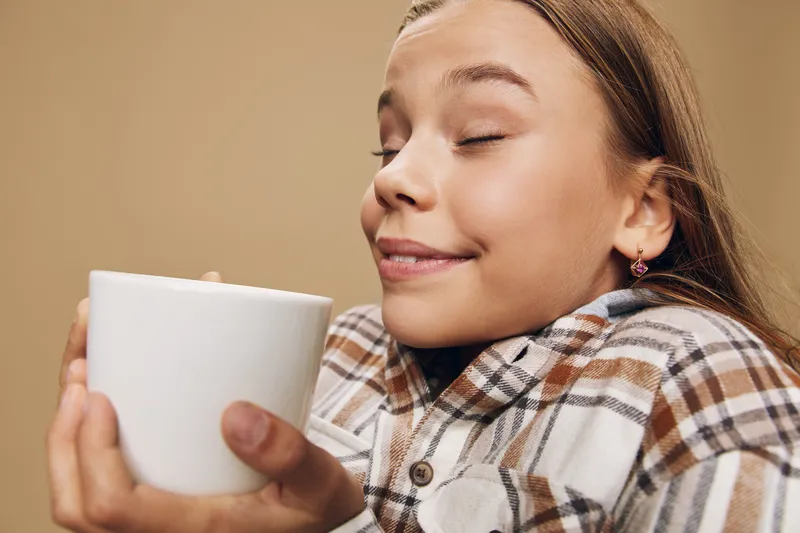 Woman holding white cup with closed eyes and smiling face, enjoying aroma and warmth. Portrait of relaxed female in plaid shirt with cozy expression against beige background.