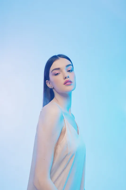 Young woman beauty fashion model poses in elegant dress against blue background. Soft lighting highlights her flawless skin and confident expression. Studio portrait concept.
