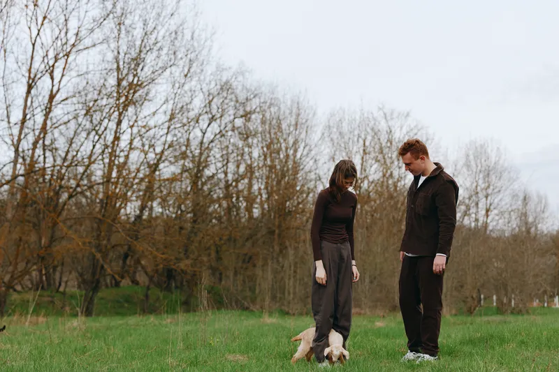 Couple standing in a grassy park, gently interacting with a small goat kid. Casual lifestyle moment, natural light, serene atmosphere, candid connection, outdoor wellness, simple joy in nature,