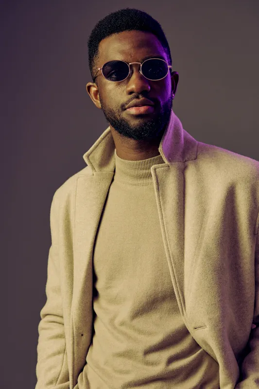 Stylish African man in a beige coat and sunglasses with a confident expression against a purple gradient background, ideal for fashion and lifestyle concepts