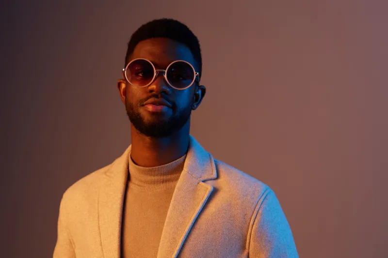 Stylish male model wearing sunglasses and a light coat, showcasing modern fashion against a colorful backdrop, exuding confidence and charisma