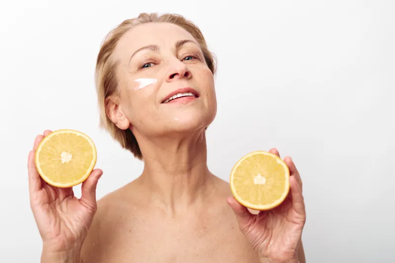 Mature woman with short hair holds two orange halves near face, smiling gently with skincare cream on cheek. Concept for natural beauty, healthy skin, wellness, and self-care.