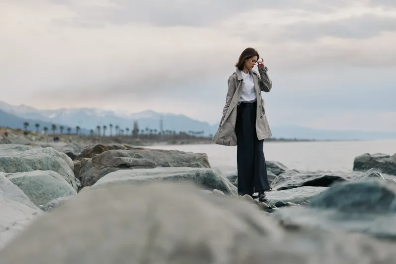 Young woman in a long coat walks on rocky beach near calm sea under cloudy sky. Mountains and palm trees are visible in the distance. Concept of solitude and nature walk.