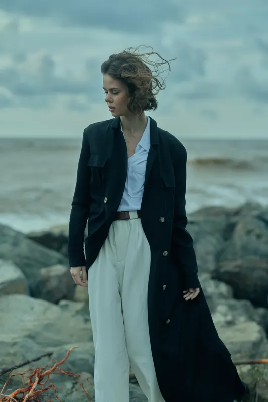 Young woman in a long dark coat and white wide-leg pants stands on a windy rocky shore, exploring fashion and sea breeze with a moody, oceanic backdrop