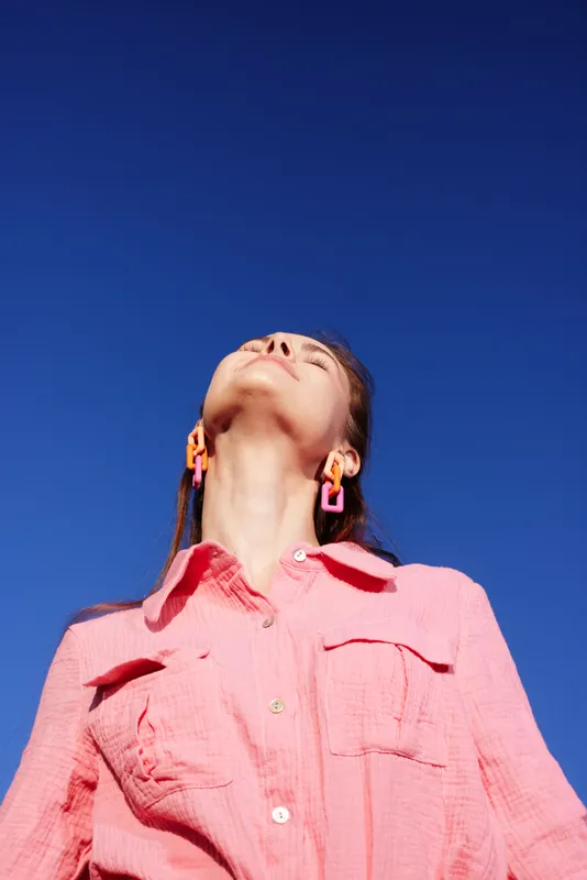 Young woman with long hair wearing pink shirt enjoying clear blue sky, showcasing positive emotions and vibrant colors, ideal for lifestyle and wellness themes