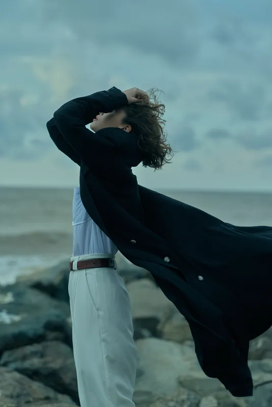 Windblown cape and flowing hair frame a figure by the sea, hands on head, ocean breeze, dramatic coastal motion, casual chic style, moody atmosphere with expansive sky and waves