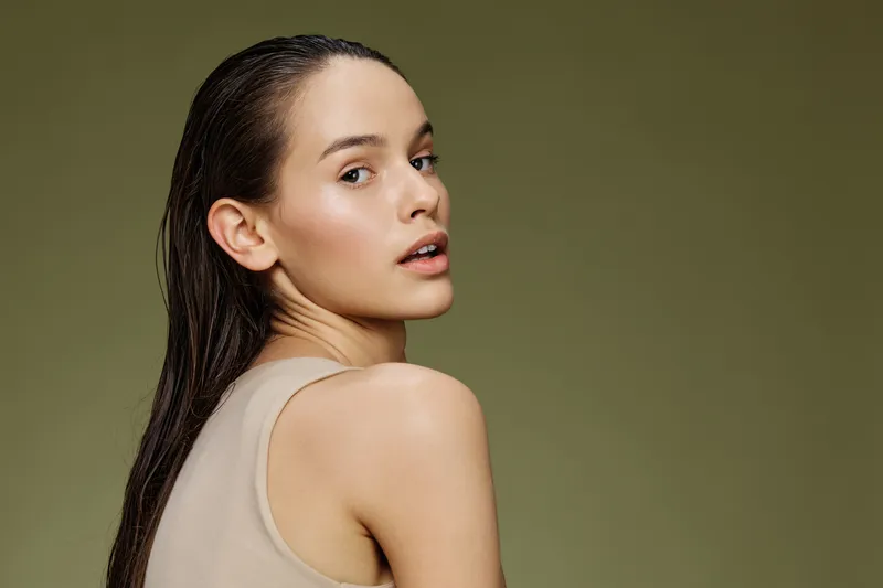 Young beautiful model with long wet hair showing natural beauty, wearing a simple tank top against a muted green background