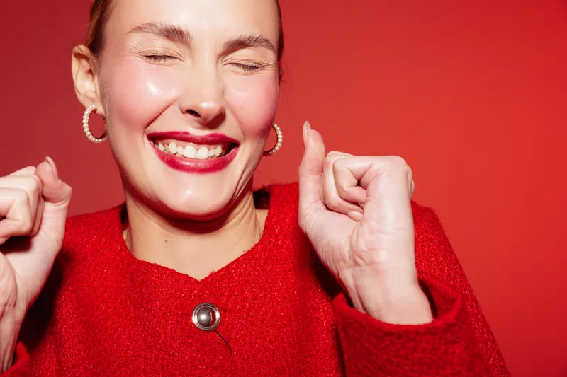 Happy woman in red sweater smiles with closed eyes and raised fists on red background. Concept of joy, celebration, positive emotions, and confident female portrait in studio lighting.