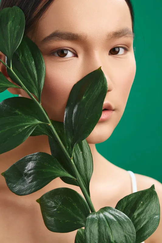 Asian woman poses in a studio portrait with lush greenery close to her face, creating a calm, natural mood and a fresh, organic atmosphere.