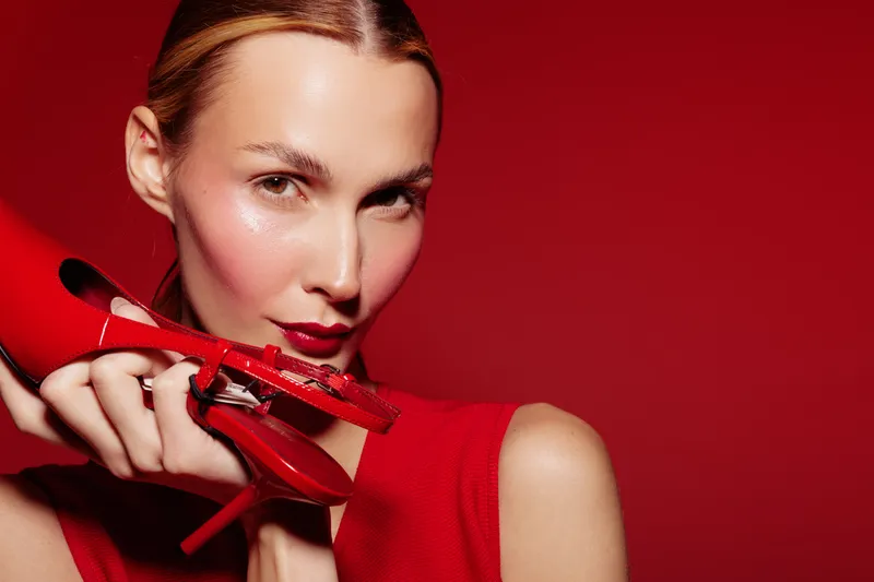 Close-up of a stylish woman holding red high heels near her face on red background. Fashion concept with bold makeup and elegant outfit in vibrant studio lighting.