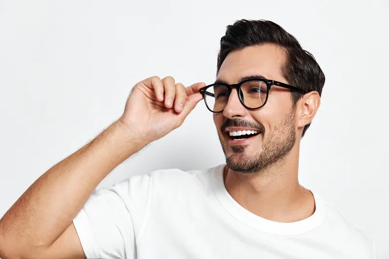 Happy smiling young man wearing glasses, holding them while looking at the camera, dressed in a casual white t-shirt against a simple light background, studio portrait.