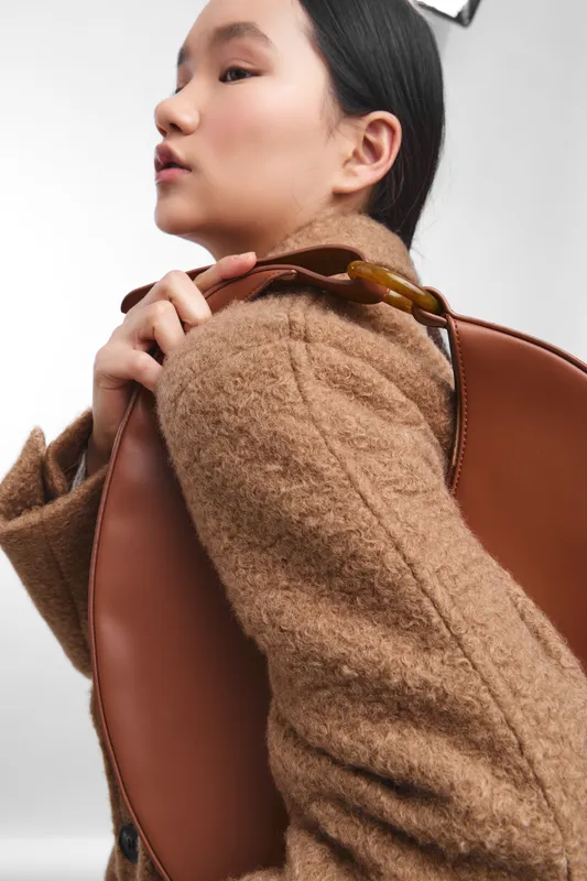 asian woman in a cozy brown coat carries a leather bag over her shoulder in a fashion studio. she wears a confident smile and a modern, chic vibe.
