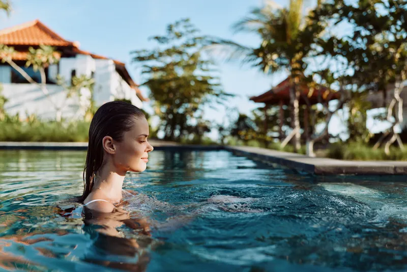 Relaxed woman in a pool at an outdoor resort, sunlit water and clear blue surroundings. Focus on leisure, wellness, and selfcare with bright warm light and a peaceful mood.