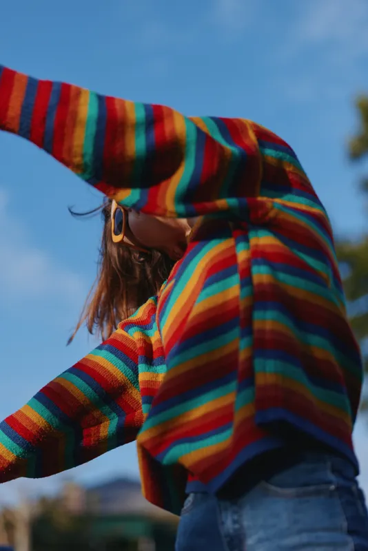 Young woman in a colorful striped sweater and retro sunglasses poses outdoors under a bright blue sky, raising an arm in a joyful, casual outfit for lifestyle fashion.