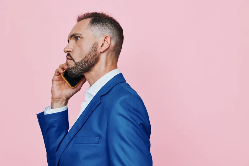 A professional man in a blue suit speaks on a mobile phone against a solid pink background, conveying focus, confidence, and business readiness in a single frame.