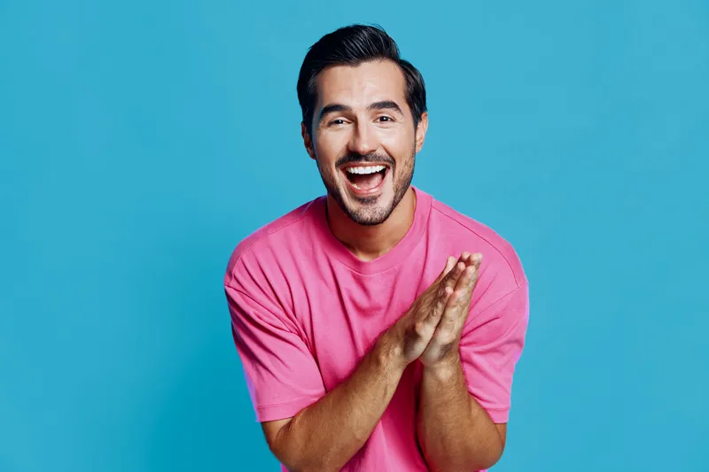 Happy smiling man wearing a pink t-shirt, looking cheerful and excited, clapping his hands in front of a solid blue background. Joyful lifestyle concept.