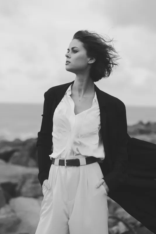Fashionable woman standing at the shore in a flowing coat and white suit, wind in hair, cinematic monochrome mood, confident stance, minimal styling, outdoor scene for editorial fashion and lifestyle