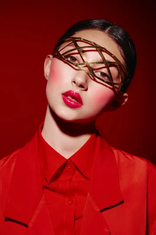 Avant-garde beauty portrait of a young woman with creative makeup and metallic face accessory in bold red fashion attire. Studio shot on red background highlighting artistic concept.