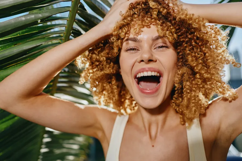 smiling woman with curly hair enjoying sunny day outdoors near tropical palm leaves, showing joyful emotion and natural beauty in warm sunlight