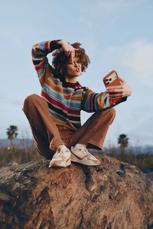 Woman lifestyle outdoors on a rock, striped sweater and casual pants, holding a smartphone for a selfie, relaxed on stone under bright sky, with palm trees, summer travel, vacation