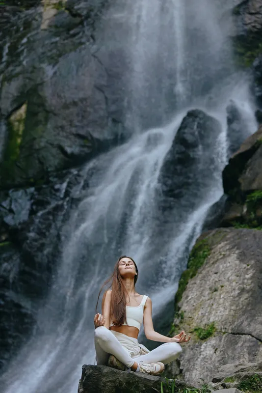Woman meditating outdoors near a waterfall, wearing white sportswear, serene expression, natural setting, peaceful moment, healthy lifestyle, mindfulness practice.
