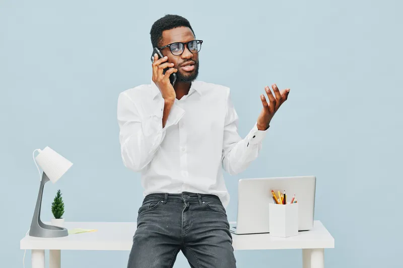 Professional man having a phone conversation in modern office environment, wearing white shirt and black jeans. Indoor workspace with desk lamp, potted plant, and laptop. People lifestyle concept.