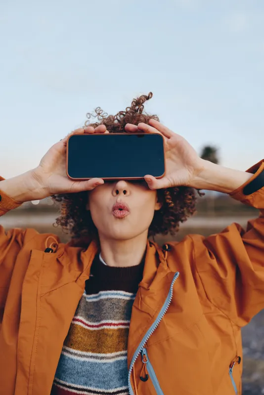 Young woman with curly hair holds a smartphone in front of her face outdoors, sky background, orange jacket, hands create playful summer modern lifestyle mood, technology concept