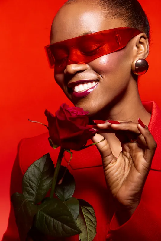 Smiling black woman with dark skin wearing red futuristic glasses and red outfit holds red rose close to her face on red background. Concept of beauty, fashion, and floral elegance.