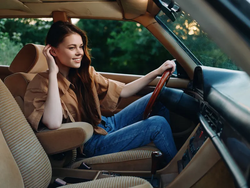 Young woman in casual attire enjoying a ride in a vintage car, conveying joy and freedom in a lush green environment.