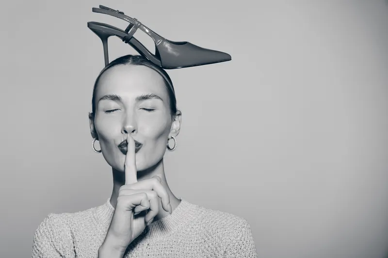 Young Caucasian woman with light skin holds finger to lips in shushing gesture while balancing high heel shoe on head. Concept for silence, fashion, and playful mood in studio.