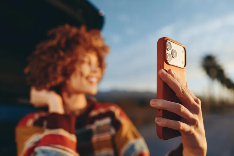 Smiling woman holds smartphone for a selfie outdoors, portrait with blurred urban street background, travel lifestyle, warm daylight and soft bokeh, casual fashion