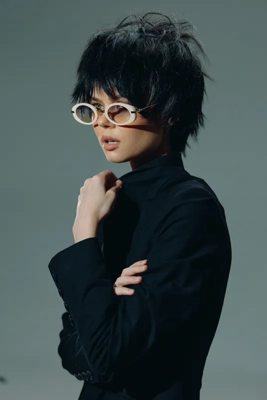 Stylish woman in black turtleneck wearing white sunglasses with curly hair and confident expression against gray background