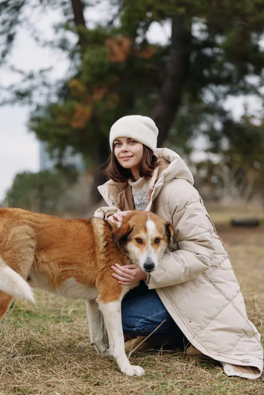 Woman in a warm beige coat kneels in a park, hugging a calm dog during autumn warmth, capturing a moment of companionship and gentle outdoor peace.