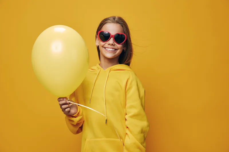 Girl wearing yellow hoodie and red heart shaped sunglasses holding yellow balloon smiling against yellow background. Portrait of happy child with casual clothes and party accessory.