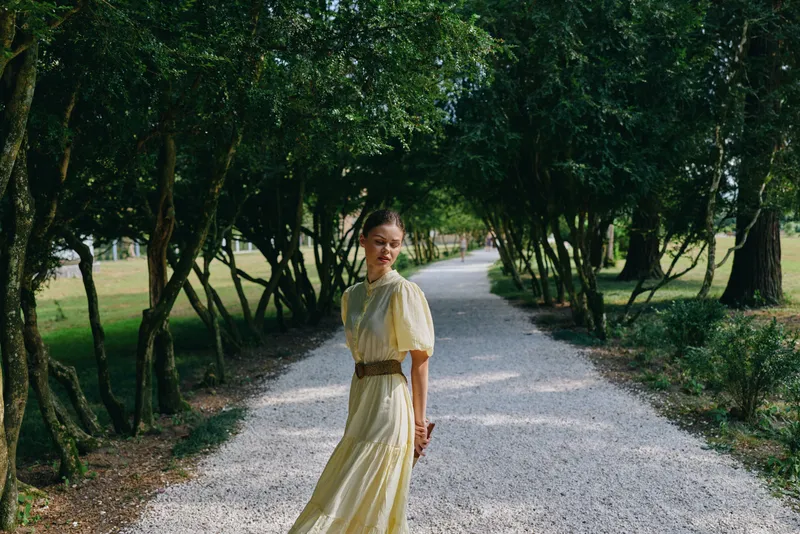 A woman in a flowing yellow dress walks along a gravel path in a sunlit park, surrounded by trees, creating a calm outdoor fashion moment.