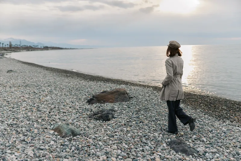 A woman in a coat and hat walks alone on a pebble beach near calm sea water at sunset. The cloudy sky and distant mountains create a peaceful, contemplative mood.