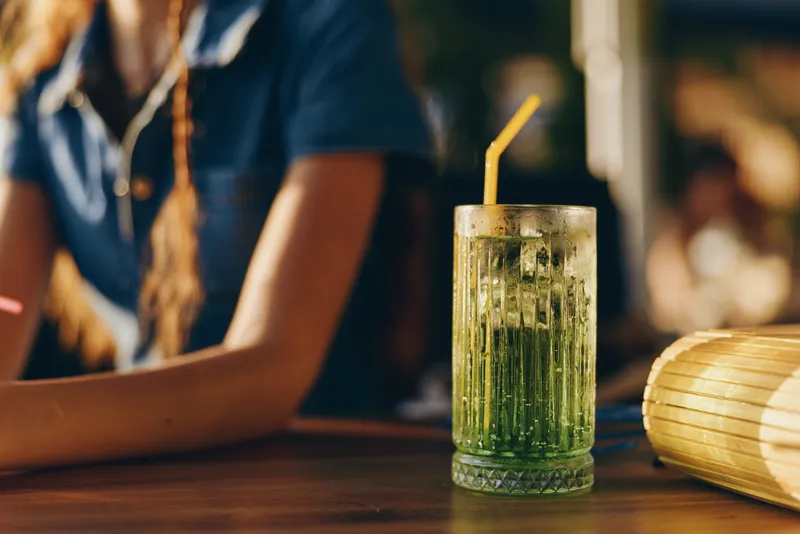 Refreshing green cocktail with ice and straw on wooden table, warm sunlight background, casual summer atmosphere and relaxed vibe.