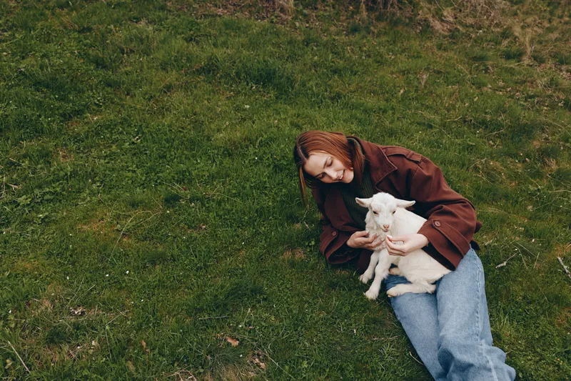 Young woman in a brown jacket lies on green grass holding a small white baby goat with a gentle and caring expression in a natural outdoor setting.