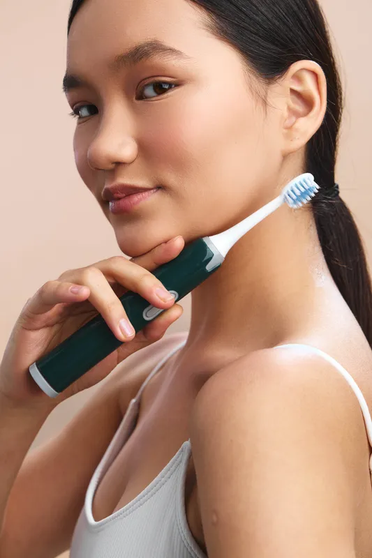 asian woman with a light top poses close to the camera, holding a toothbrush near her face as she demonstrates dental care in a clean, confident lifestyle setting.