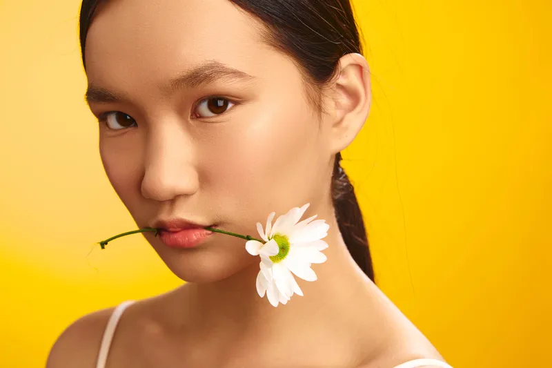 asian woman with a delicate flower held between her lips stands against a bright yellow backdrop, offering a calm, artistic studio portrait with refined beauty and subtle mood.