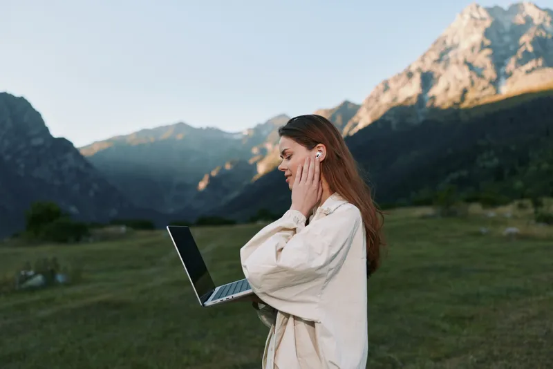 A focused professional stands outdoors with a laptop in hand, surrounded by rugged mountains and green fields, conveying remote work, nature, and concentration in a tranquil setting.