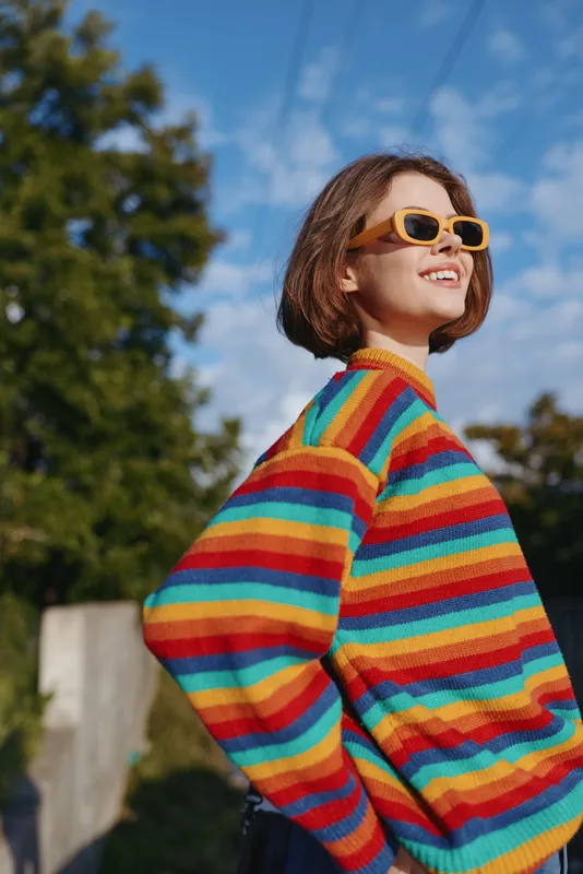 Young woman in a rainbow sweater and yellow sunglasses smiling outdoors under blue sky, casual outfit and short bob hairstyle, sunlit fashion portrait with joyful expression.