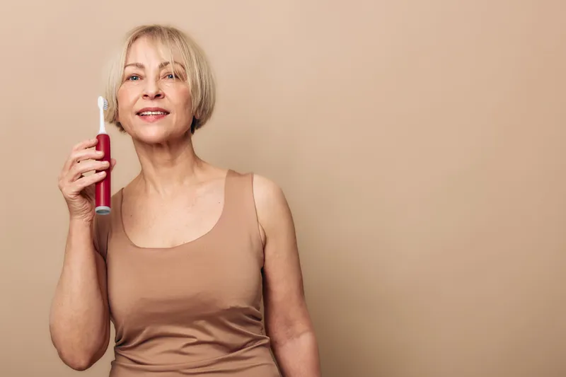 Mature woman with short blonde hair holds electric toothbrush smiling against beige background. Concept of dental hygiene, oral care, and healthy lifestyle for seniors.