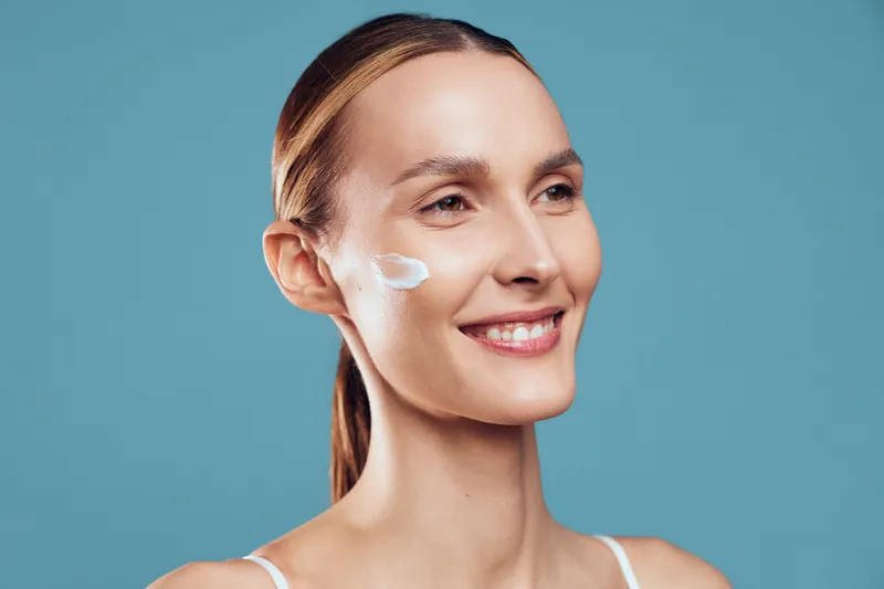A smiling woman with clear skin applies moisturizing cream on her cheek against a blue background. Concept for skincare, beauty routine, and healthy lifestyle.