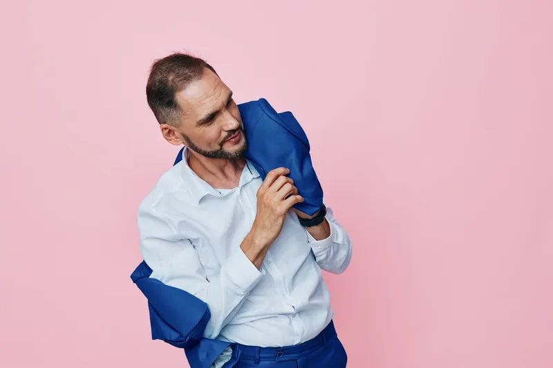 A professional man in business attire stands against a vibrant pink background, removing a blue jacket as he projects a thoughtful mood and confident demeanor.