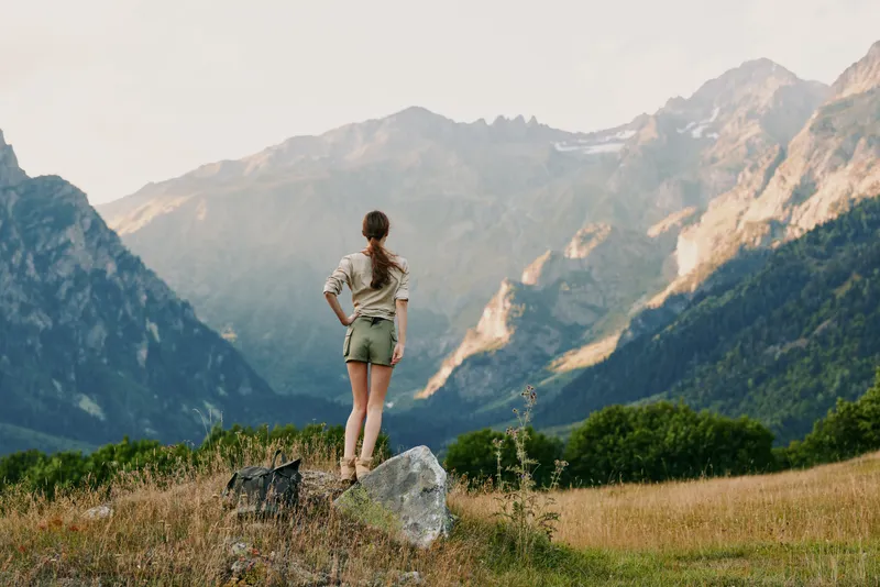 A young woman stands on a rocky outcrop in a sunlit mountain valley, hands on hips, surveying distant peaks, ready for a hike, evoking freedom and adventure.