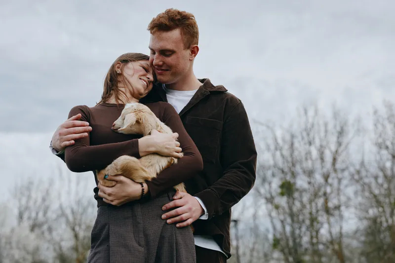 A couple outdoors cuddle a goat kid, feeling warmth and bonding in a rustic countryside setting. Lifestyle moment showcasing family affection, gentle animal companionship, and everyday joy for